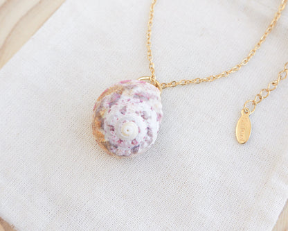 Sunny Pink White Orange Magus Top Shell Gold Necklace on display side view. A gold-coloured chain necklace with a seashell pendant. The pendant is a mix of pink, white, and gold. The necklace is displayed on a white cloth.