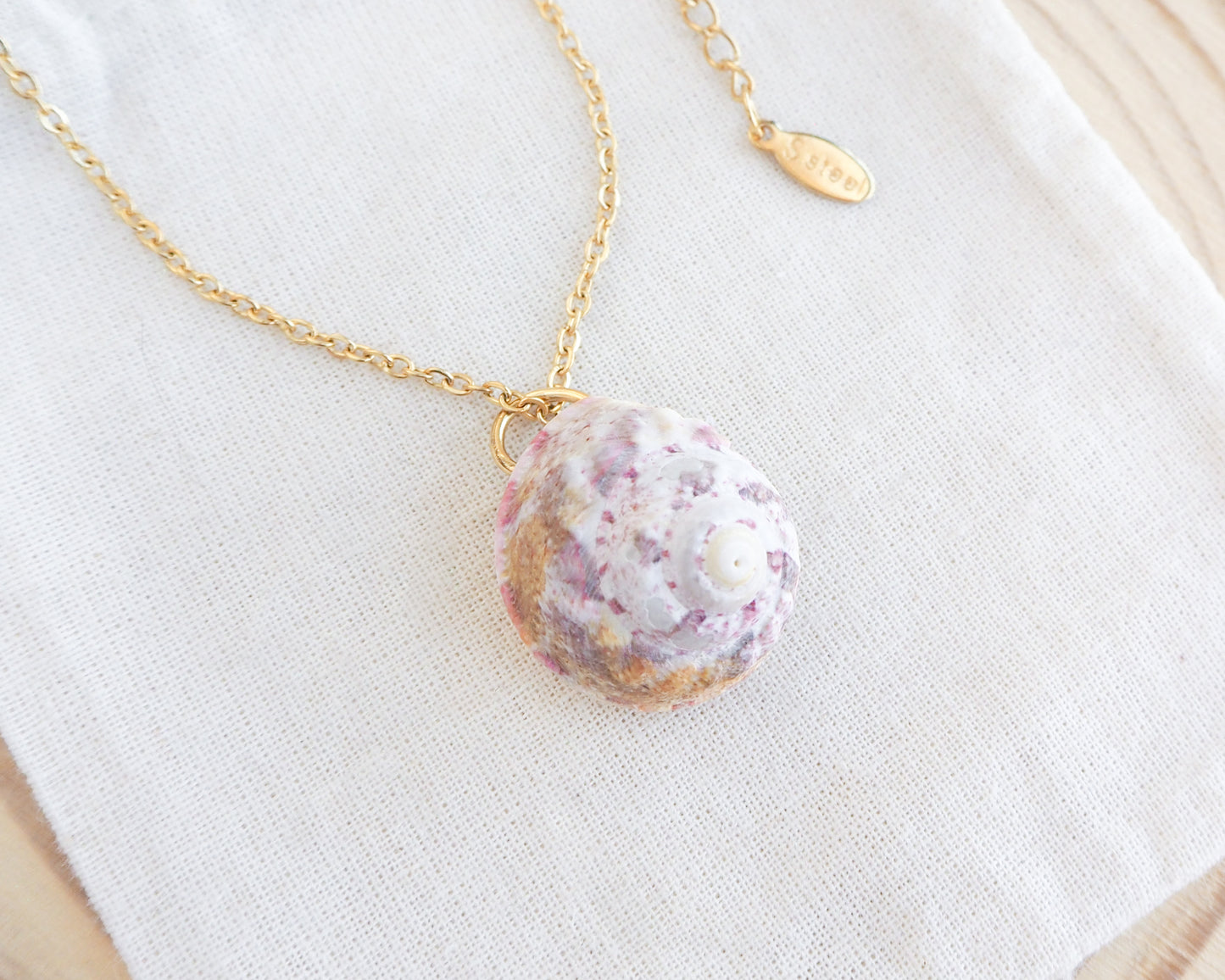 Sunny Pink White Orange Magus Top Shell Gold Necklace on display. A gold chain necklace featuring a seashell pendant. The shell is coloured with shades of white, pink, and orange. The necklace is presented on a white cloth.