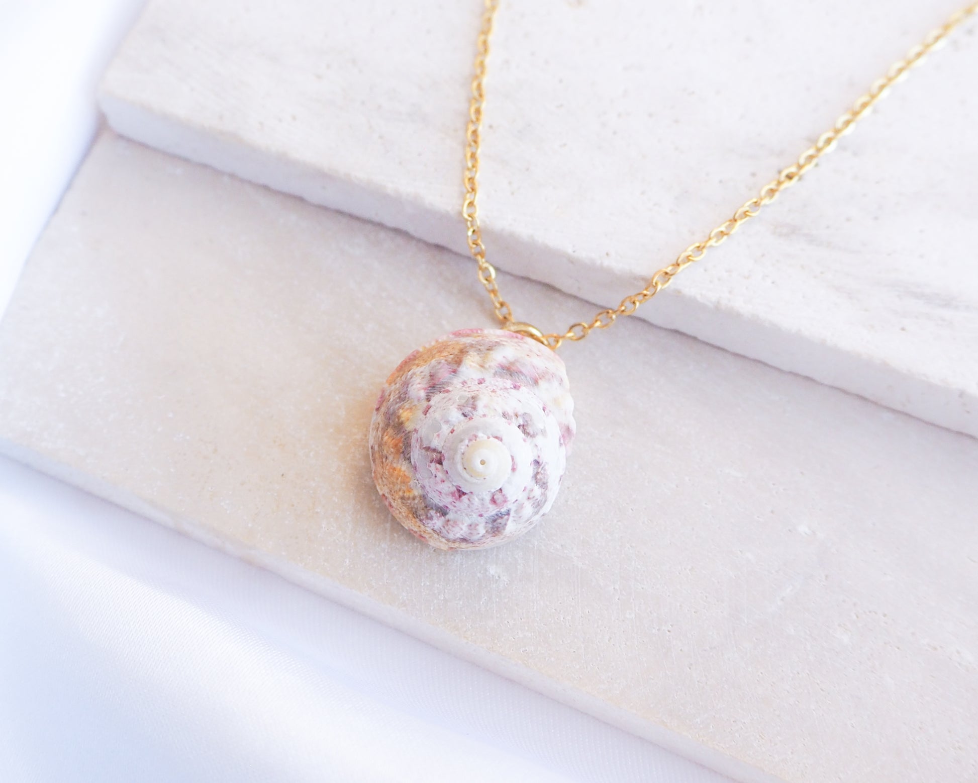 Sunny Pink White Orange Magus Top Shell Gold Necklace on stone display. A gold-coloured chain necklace with a seashell pendant. The pendant is an oval-shaped seashell with a spiral pattern, in shades of pink, white, and orange. The necklace is displayed on a white surface.