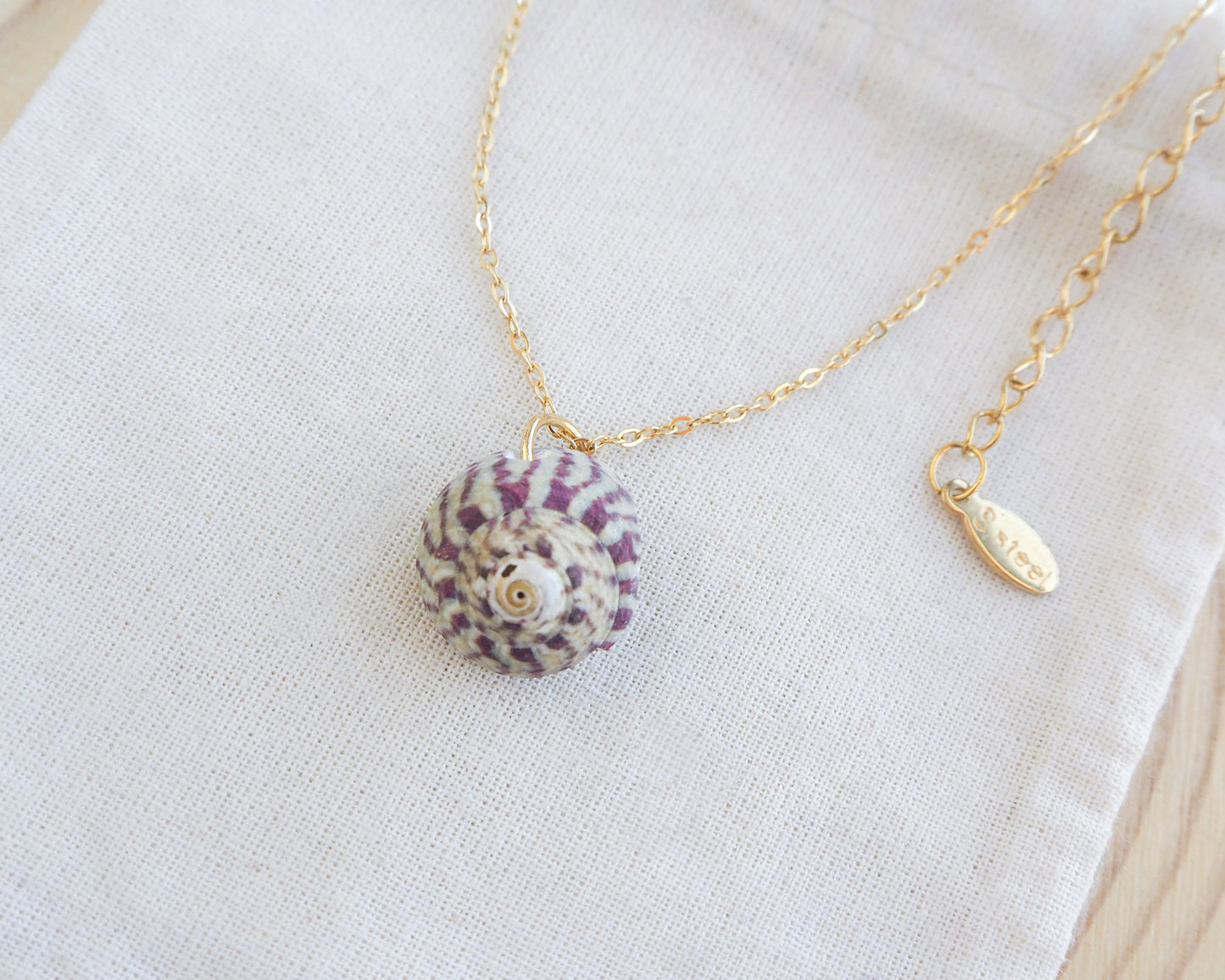 Necklace with a shell pendant on a white fabric background.  Fuchsia Green Striped Top Shell Necklace on display, with gold stainless steel chain. Real shell necklace from Portugal. A gold chain necklace featuring a seashell pendant. The shell has a spiral design in purple and white. The necklace rests on a white cloth, showcasing the jewellery's details.