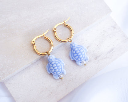 Gold hoop earrings with blue turtle charms on a white surface. Gold hoop earrings with blue turtle charms on a white background