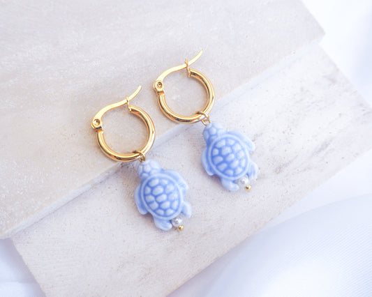 Gold hoop earrings with blue turtle charms on a white surface. Gold hoop earrings with blue turtle charms on a white background