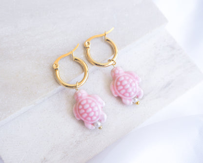 Pair of gold hoop earrings with pink turtle-shaped charms on a white background. Soft Pink Ceramic Turtle earrings with gold hoops on a light background