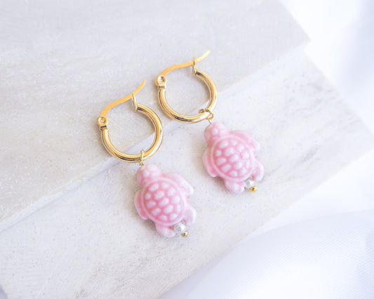 Pair of gold hoop earrings with pink turtle-shaped charms on a white background. Soft Pink Ceramic Turtle earrings with gold hoops on a light background