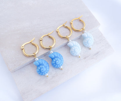 Gold hoop earrings with blue seashell-like charms on a light gray surface. Four pairs of gold-coloured hoop earrings with light blue seashell charms. Each earring has a small white bead and a gold-coloured accent. The earrings are displayed on a light grey surface.