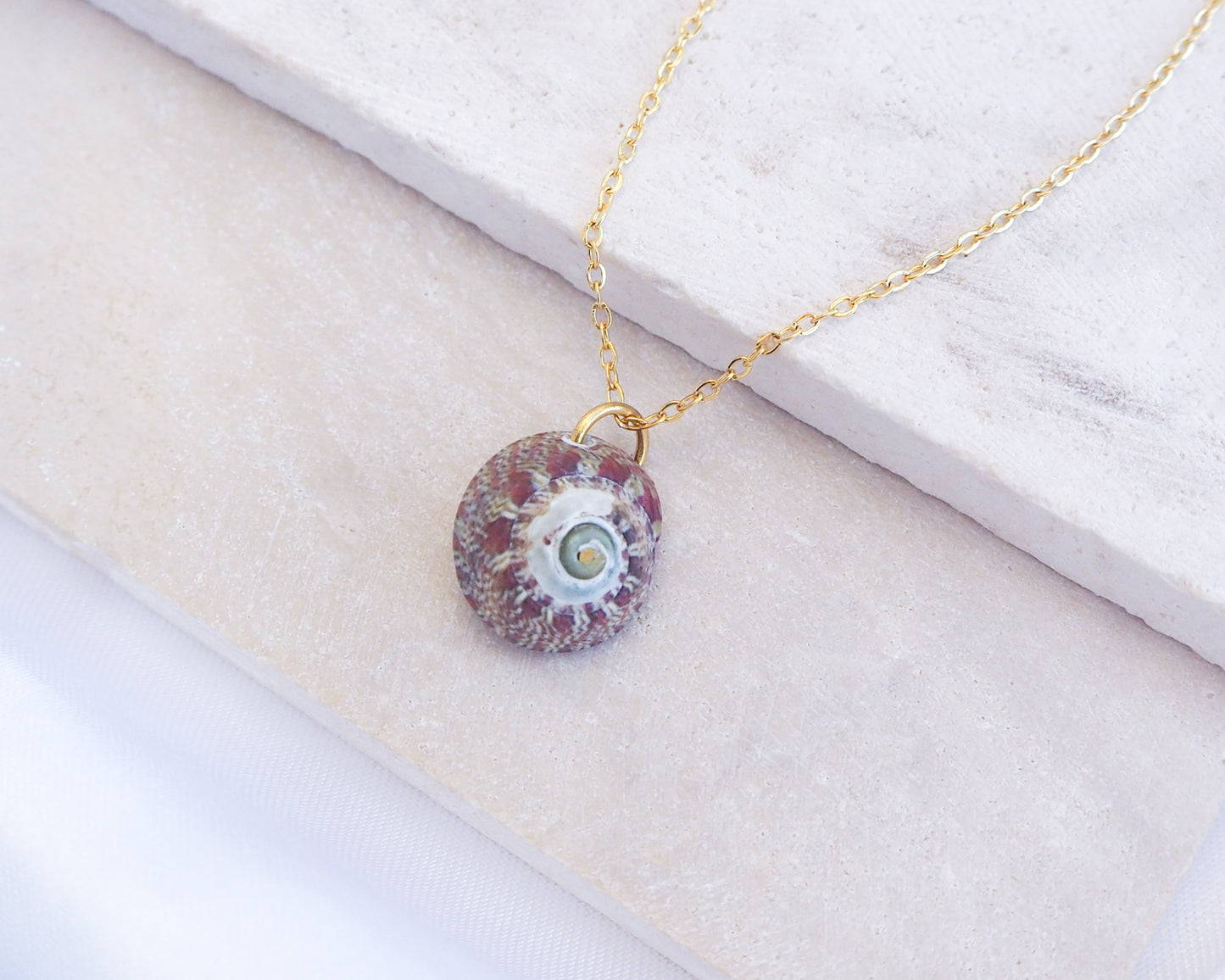 Necklace with a round pendant on a white surface. Bordeaux Green Patterned Flat Top Shell Gold Necklace on stone displayed, Flat Topshell from Portugal