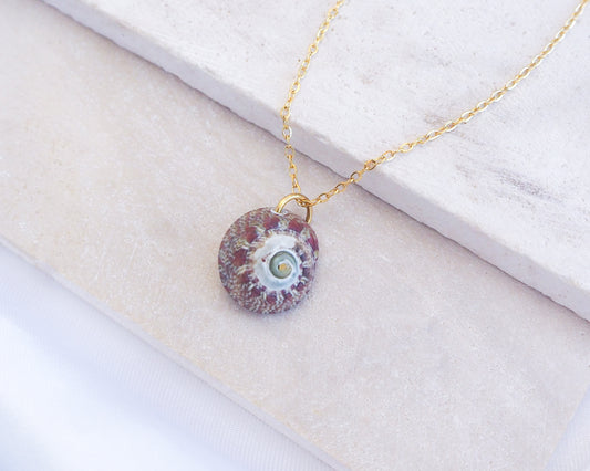 Necklace with a round pendant on a white surface. Bordeaux Green Patterned Flat Top Shell Gold Necklace on stone displayed, Flat Topshell from Portugal