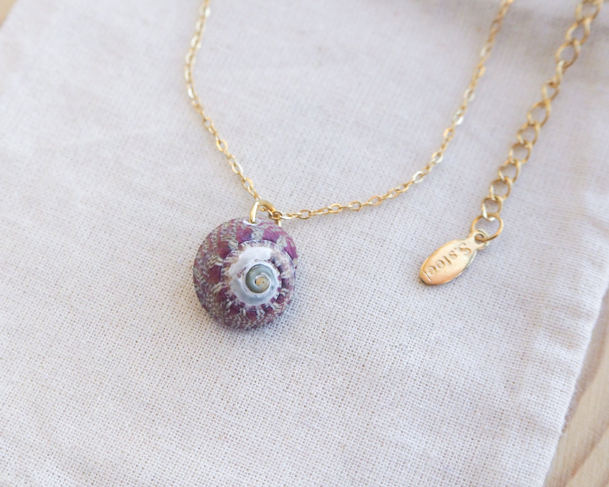Necklace with a purple pendant on a light fabric background.  Gold Bordeaux Green Patterned Flat Top Shell Necklace on display close up, Flat Topshell from Portugal