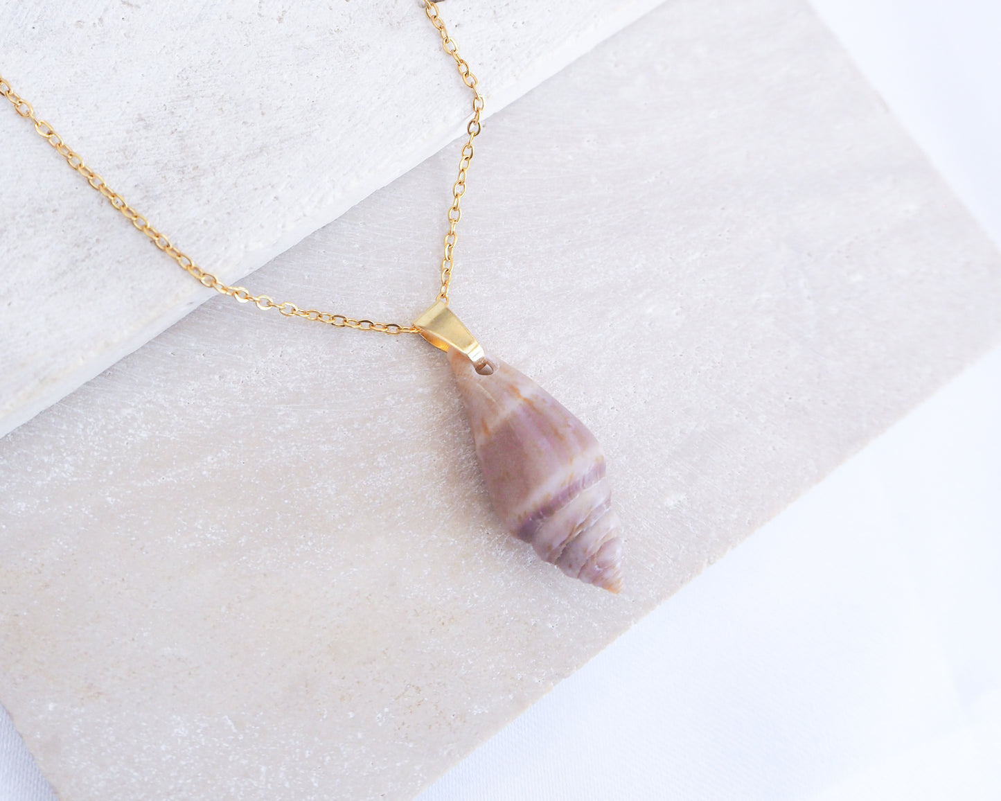 Shell pendant necklace on a gold chain against a white background. A gold-coloured chain necklace featuring a pendant made from a purple and white seashell. The seashell pendant is elongated and has a spiral design. The necklace is displayed on a white surface.