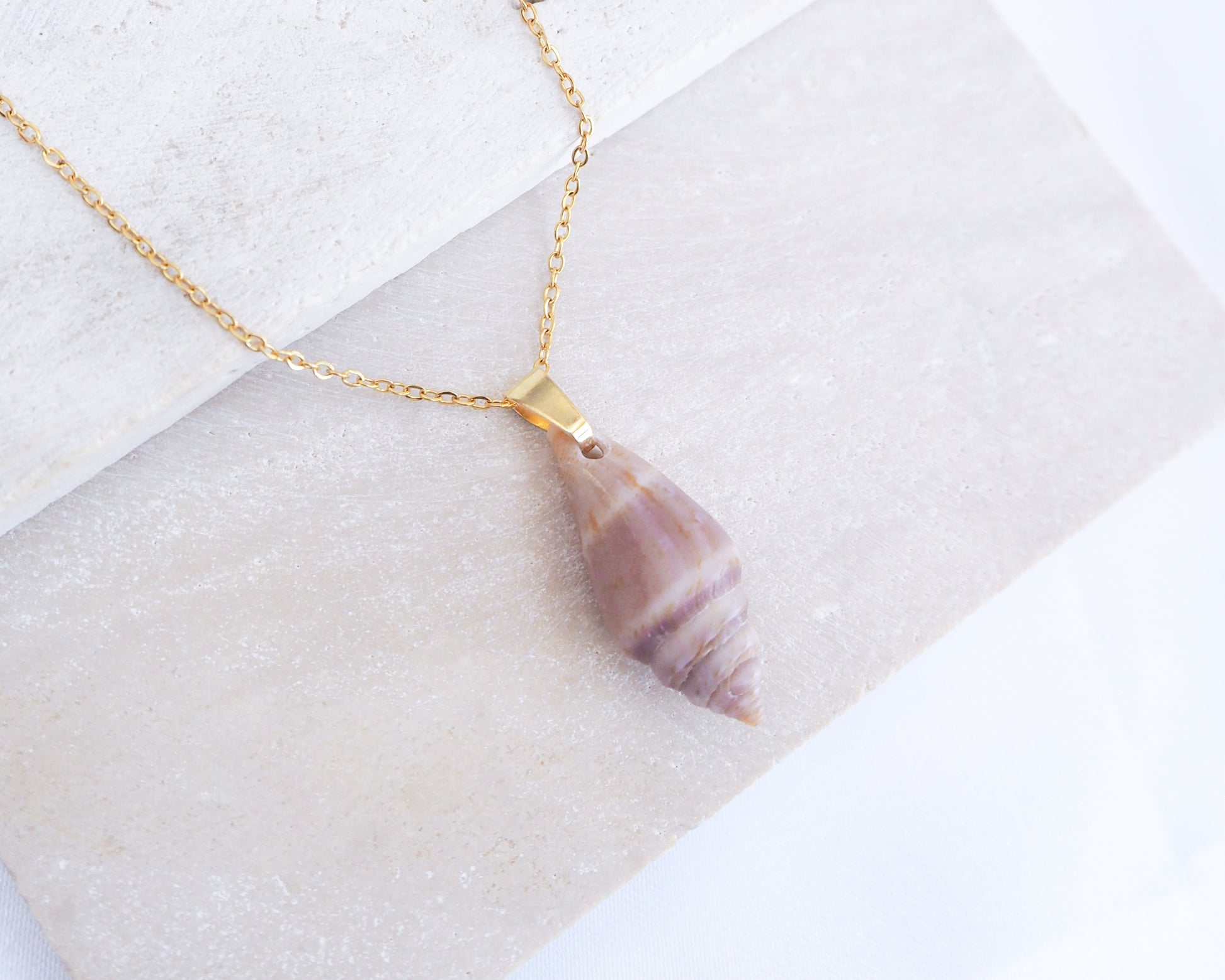 Shell pendant necklace on a gold chain against a white background. A gold-coloured chain necklace featuring a pendant made from a purple and white seashell. The seashell pendant is elongated and has a spiral design. The necklace is displayed on a white surface.
