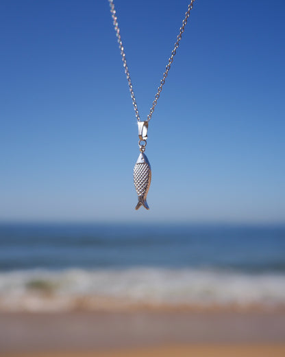 Silver fish pendant necklace against a beach backdrop with blue sky and ocean.