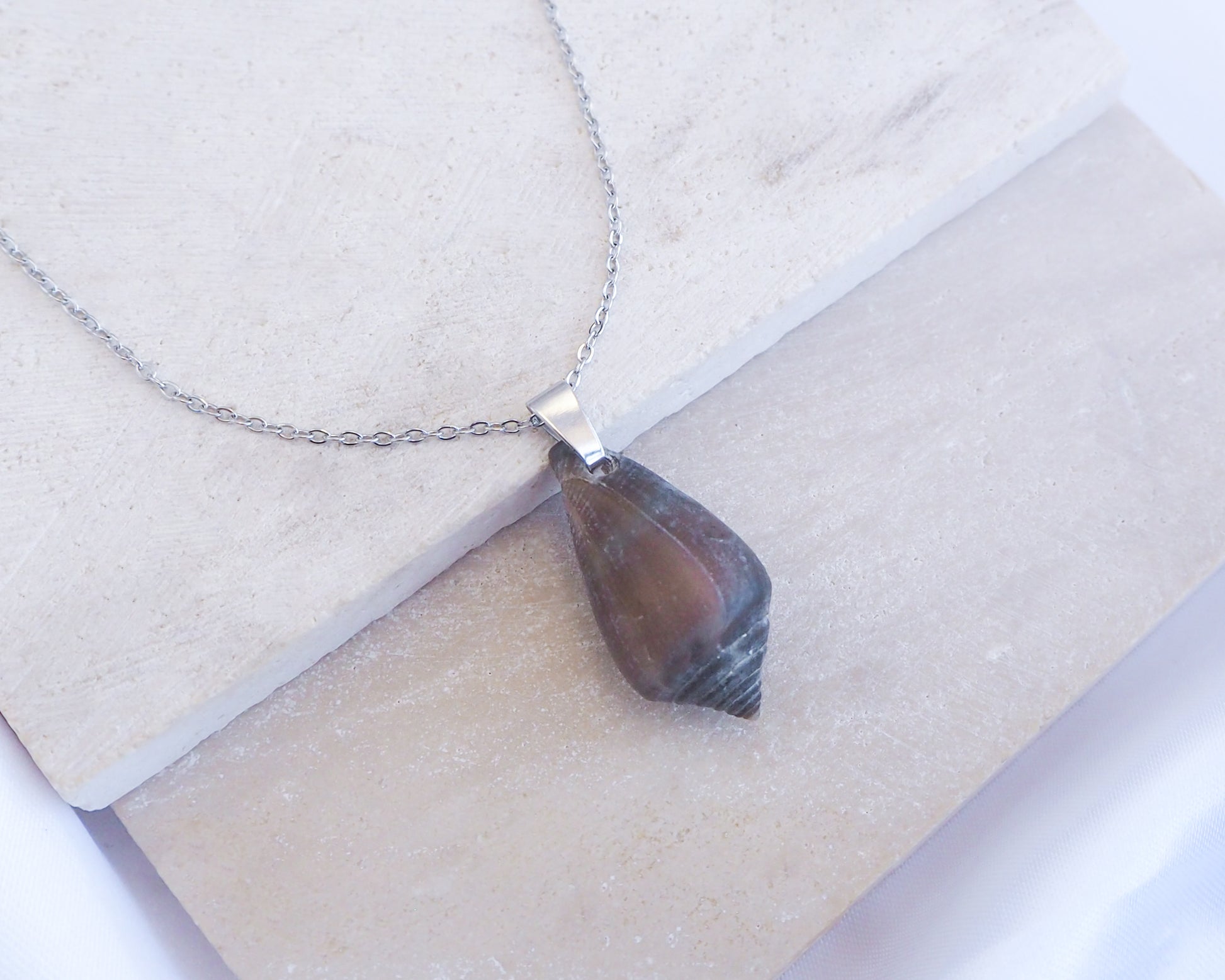 Necklace with a shell pendant on a light background. Dark Mediterranean Cone Shell Silver Necklace displayed on stone, Silver stainless steel necklace with dark cone shell from Portugal, Dark Cone Shell pendant