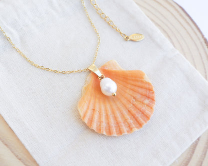 Necklace with a shell and pearl pendant on a white fabric background. Close up of Coral Orange Scallop Shell Pearl Gold Necklace II on display, Pilgrims Scallop Shell from Portugal with freshwater pearl and gold chain. 