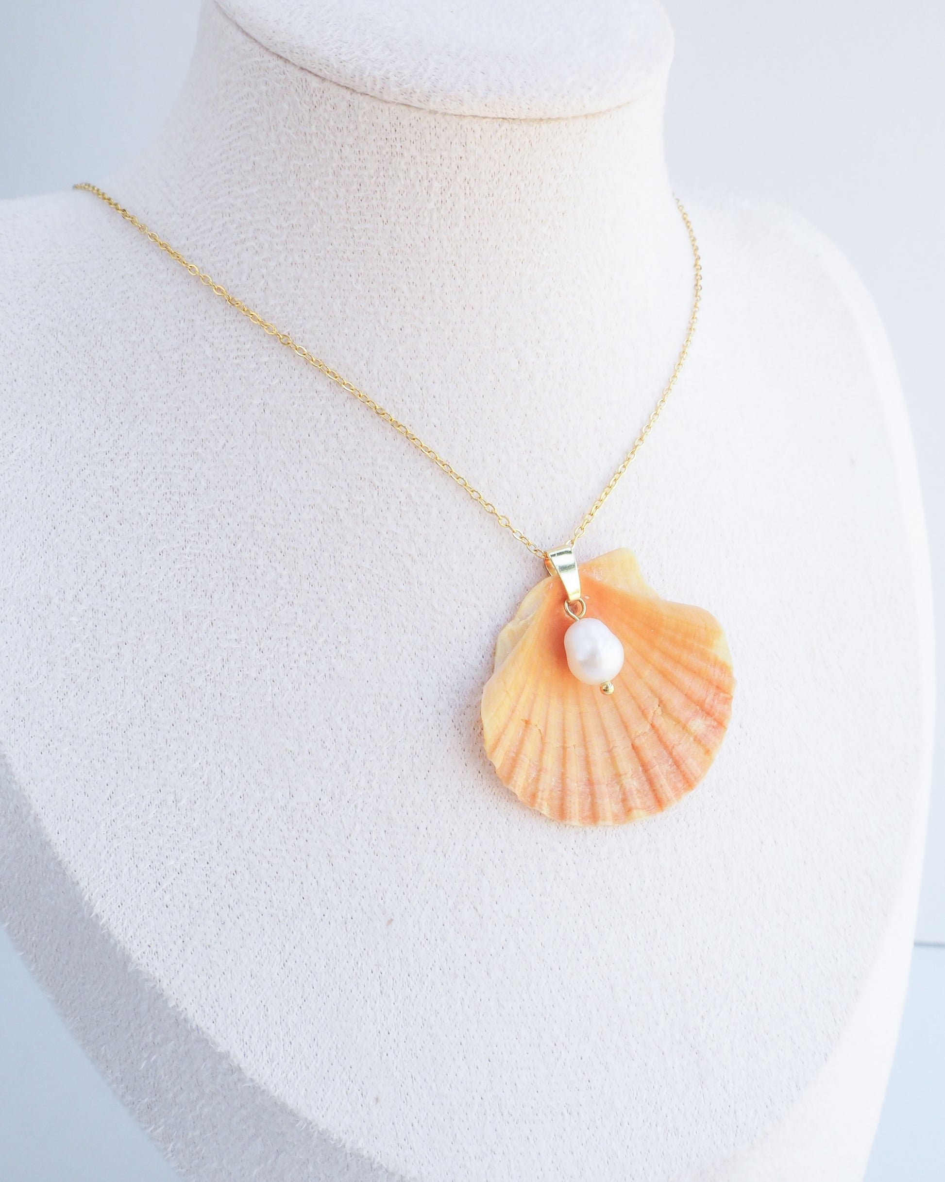Necklace with a shell pendant and pearl on a mannequin bust. Display wearing Coral Orange Scallop Shell Pearl Gold Necklace II on stone displayed, Pilgrims Scallop Shell from Portugal with freshwater pearl and gold chain. 