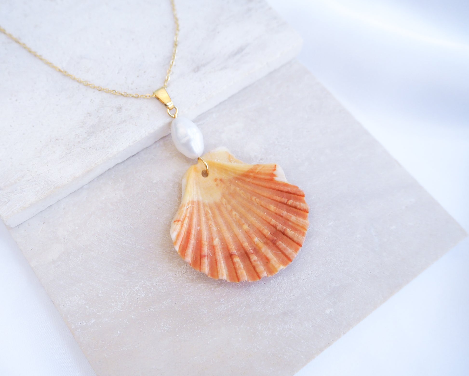 Shell pendant necklace with pearl on a white background. Coral Orange Scallop Shell Pearl Gold Necklace IV on stone displayed, Pilgrims Scallop Shell from Portugal with freshwater pearl and gold chain. 