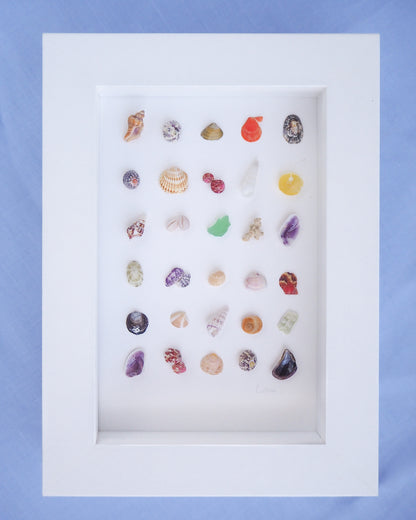 Assorted colorful stones displayed in a white frame against a blue background. Coastal artwork Seashell Mini Mix featuring 30 tiny beach treasures and shells handpicked from Portugal set in a white glass frame box. Coastal Beach Art for nightstand and bookshelf. seabylou, SEA by LOU