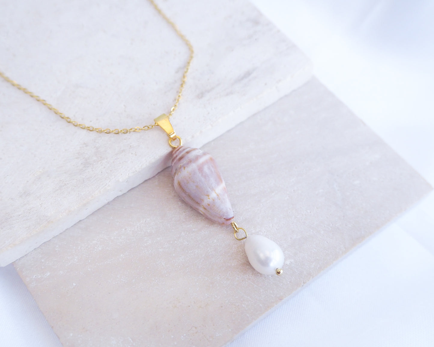 Necklace with a marbled pendant on a textured surface. Close up of Model wearing Mediterranean Cone Shell Pearl Necklace Reversed from Portugal on stone, handpicked cone shell from Algarve, Freshwater pearl and 18k gold stainless steel chain