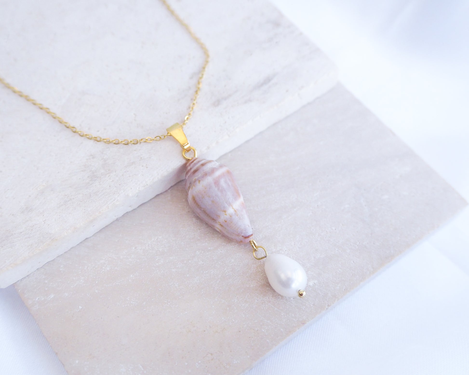 Necklace with a marbled pendant on a textured surface. Close up of Model wearing Mediterranean Cone Shell Pearl Necklace Reversed from Portugal on stone, handpicked cone shell from Algarve, Freshwater pearl and 18k gold stainless steel chain