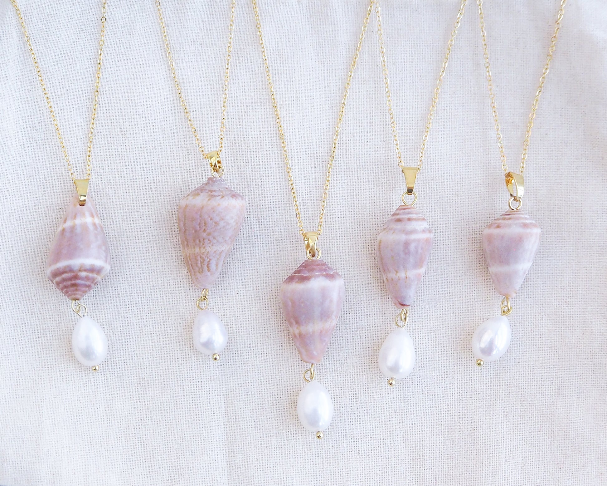 Five pearl necklaces with shell pendants on a white background. Mix of Mediterranean Cone Shell Pearl Necklaces Reversed from Portugal displayed, handpicked cone shell from Algarve, Freshwater pearl and 18k gold stainless steel chain