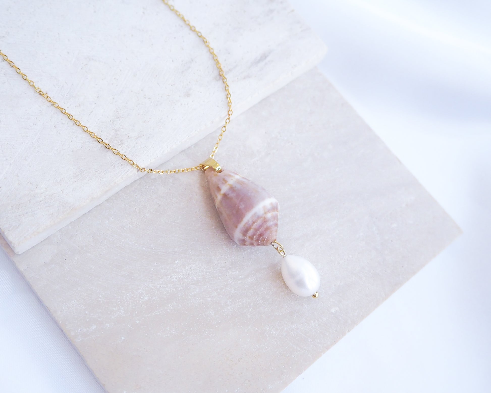 Necklace with shell and pearl pendant on a white surface. Mediterranean Cone Shell Pearl Necklace Reversed from Portugal on stone displayed, handpicked cone shell from Algarve, Freshwater pearl and 18k gold stainless steel chain
