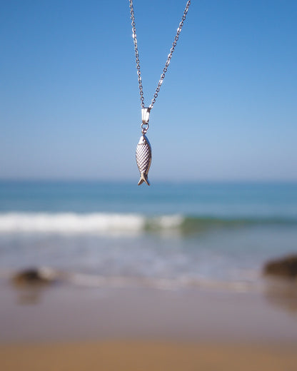 Silver fish pendant necklace against a beach backdrop with blue sky and ocean.