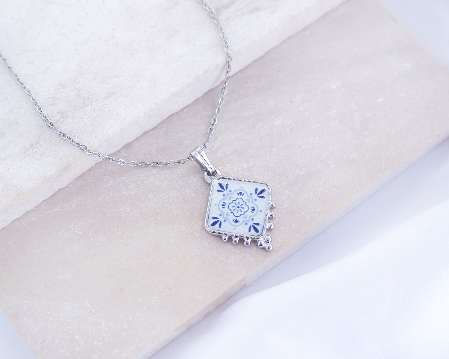 Close up of Portuguese Tile Necklace Blue Blossom Silver on stone displayed. Made with silver stainless steel and white blue floral pendant inspired by the Portuguese Azulejo design pattern. 