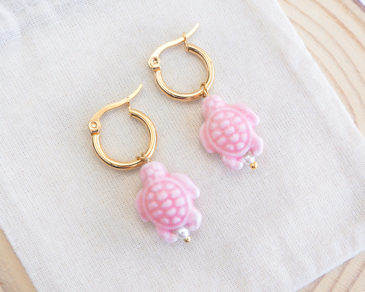 Soft Pink Ceramic Turtle earrings on gold hoops displayed on a white cloth.