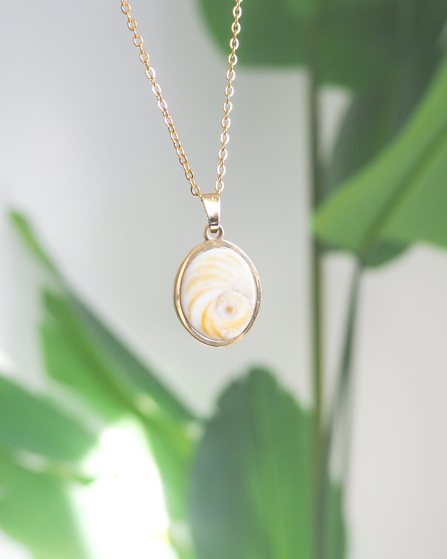 Front view of Soft Yellow Shiva Eye Necklace with green plant background. Light Eye of Saint handpicked from Portugal, 18k gold stainless steel shiva eye pendant necklace, Saint Lucy Eye, operculum pendant