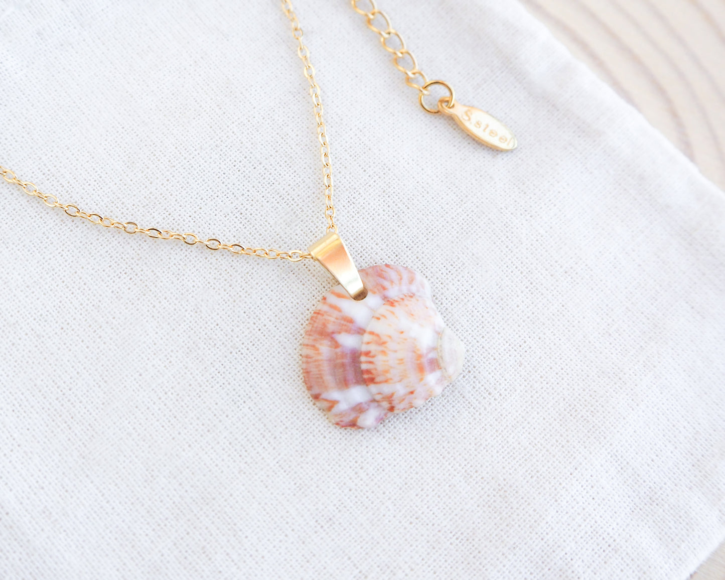 Strawberry venus seashell pendant. The pendant is a natural seashell with red and white hues with a unique marble pattern. The chain and clasp are 18k gold stainless steel. The necklace is displayed on a white cloth. The necklace has a delicate chain and an adjustable clasp. The pendant is approximately 2.5 cm in length.