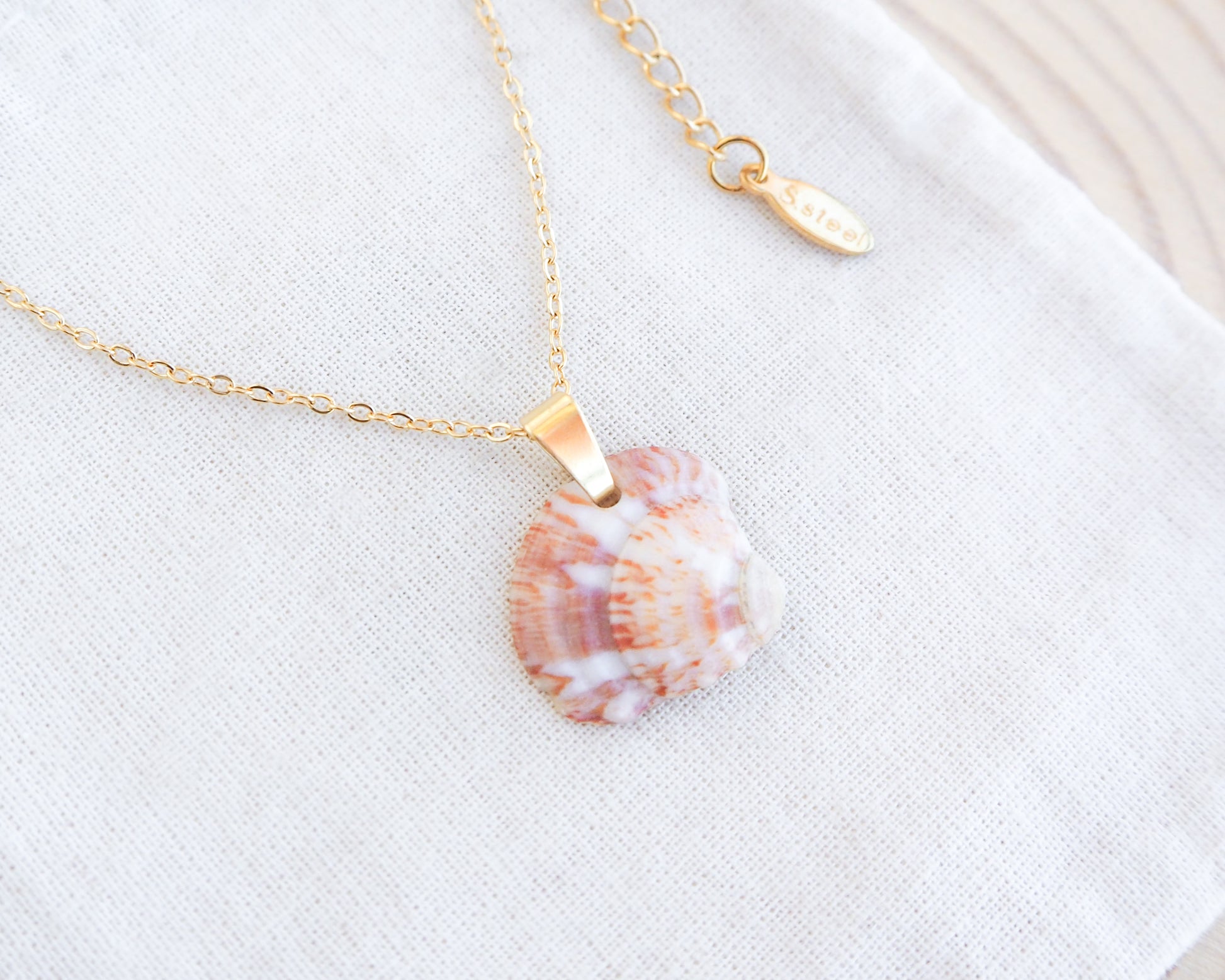 Strawberry venus seashell pendant. The pendant is a natural seashell with red and white hues with a unique marble pattern. The chain and clasp are 18k gold stainless steel. The necklace is displayed on a white cloth. The necklace has a delicate chain and an adjustable clasp. The pendant is approximately 2.5 cm in length.