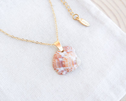 Strawberry venus seashell pendant. The pendant is a natural seashell with red and white hues with a unique marble pattern. The chain and clasp are 18k gold stainless steel. The necklace is displayed on a white cloth. The necklace has a delicate chain and an adjustable clasp. The pendant is approximately 2.5 cm in length.