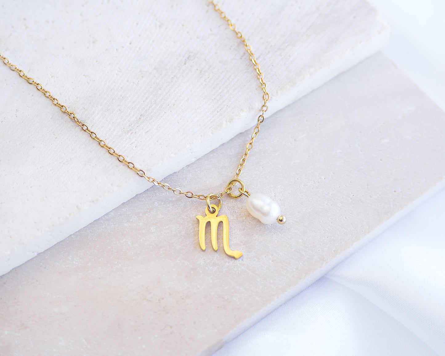 Gold necklace with a zodiac symbol and pearl pendant on a white background. Gold Scorpio Zodiac Pearl Necklace on stone displayed, made with 18k gold stainless steel and white freshwater pearl 